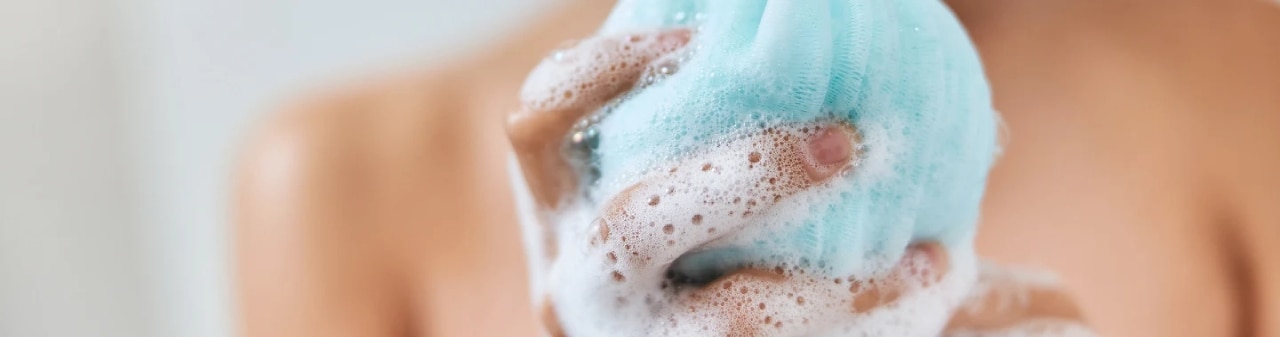 rutina-para-piel-suave mujer sonriente con un paño exfoliante espumoso en la mano tomando la ducha en casa